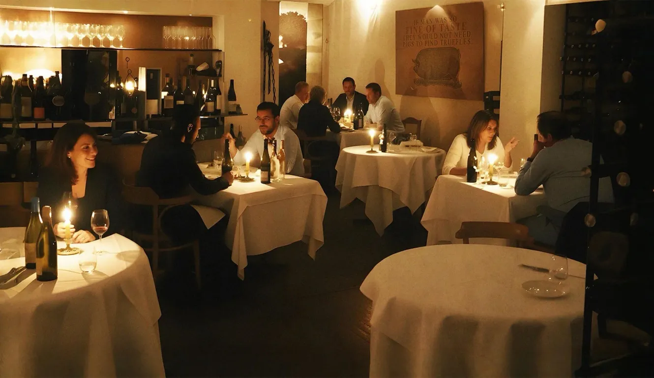 Sfeervolle restaurantsetting met tafels, wijnflessen, kaarslicht en gasten.